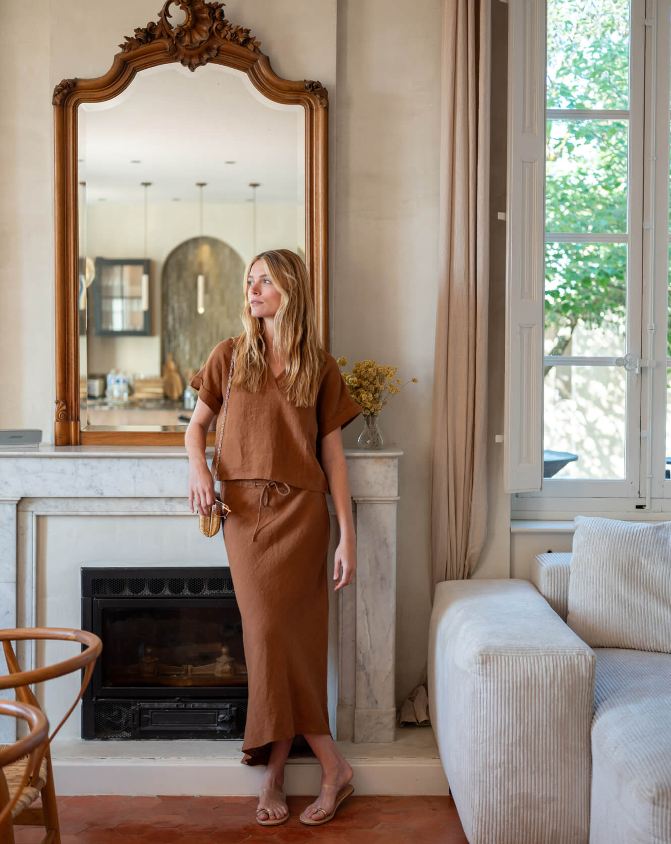 Woman in a brown outfit standing in a living room with a large mirror and fireplace.