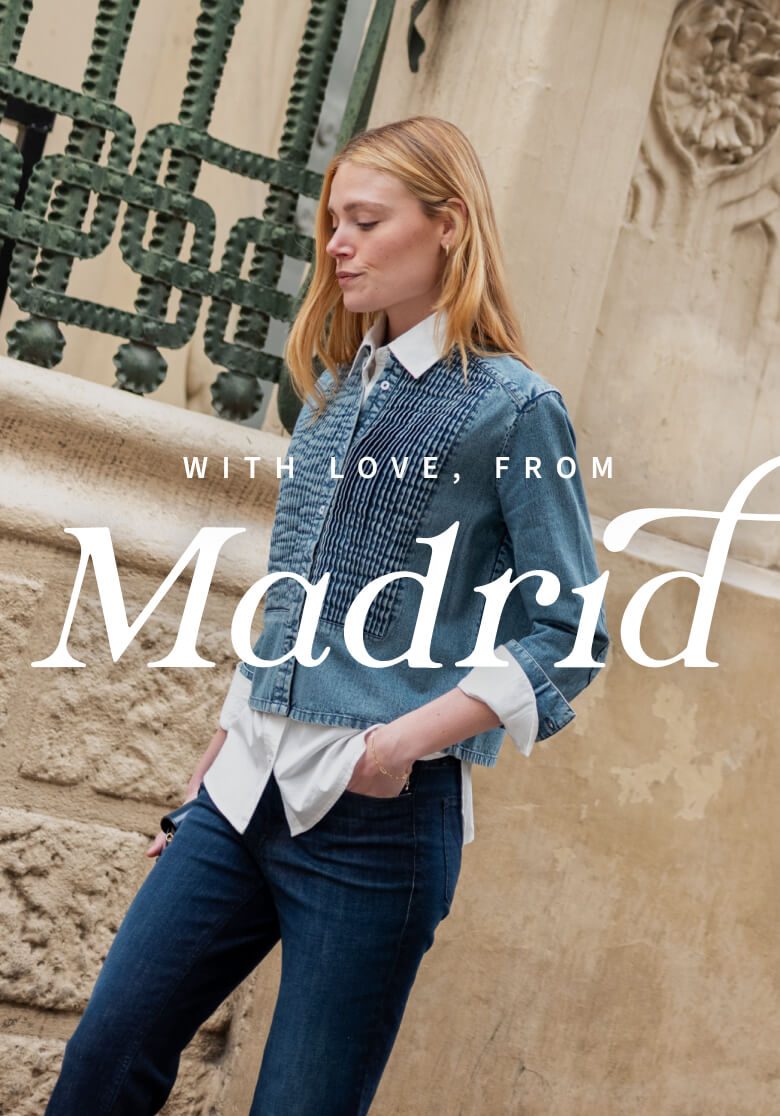 A woman wearing a blue denim-style jacket over a white button-down shirt and denim jeans, standing in front of a decorative wrought iron gate and stone wall. Text on image: WITH LOVE, FROM Madrid