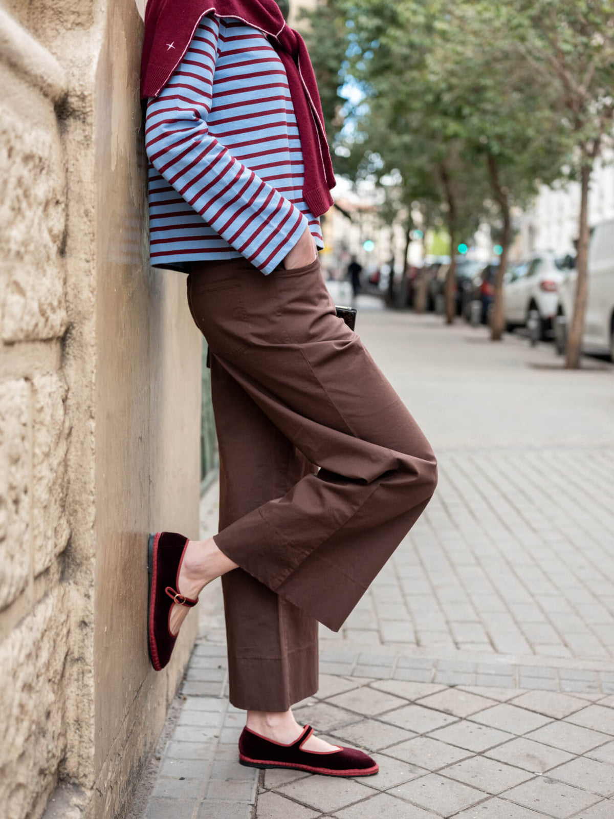 A woman in a striped top, a deep red wrap, and wide-leg brown trousers stands against a stone wall on a city street in Madrid, wearing flat mules.