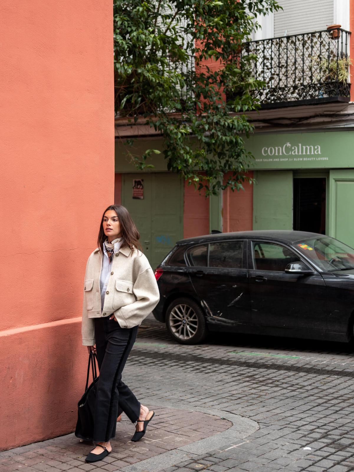 A woman wearing a light beige knit cardigan and light blue shirt walks along a cobblestone street past a terracotta-colored building and a parked black car. conCalma