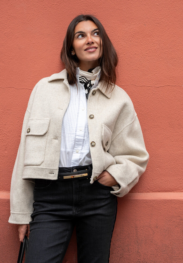 A woman smiles while leaning against a textured terracotta wall, wearing a cream oversized wool-blend utility jacket over a blue and white striped shirt with a silk scarf.
