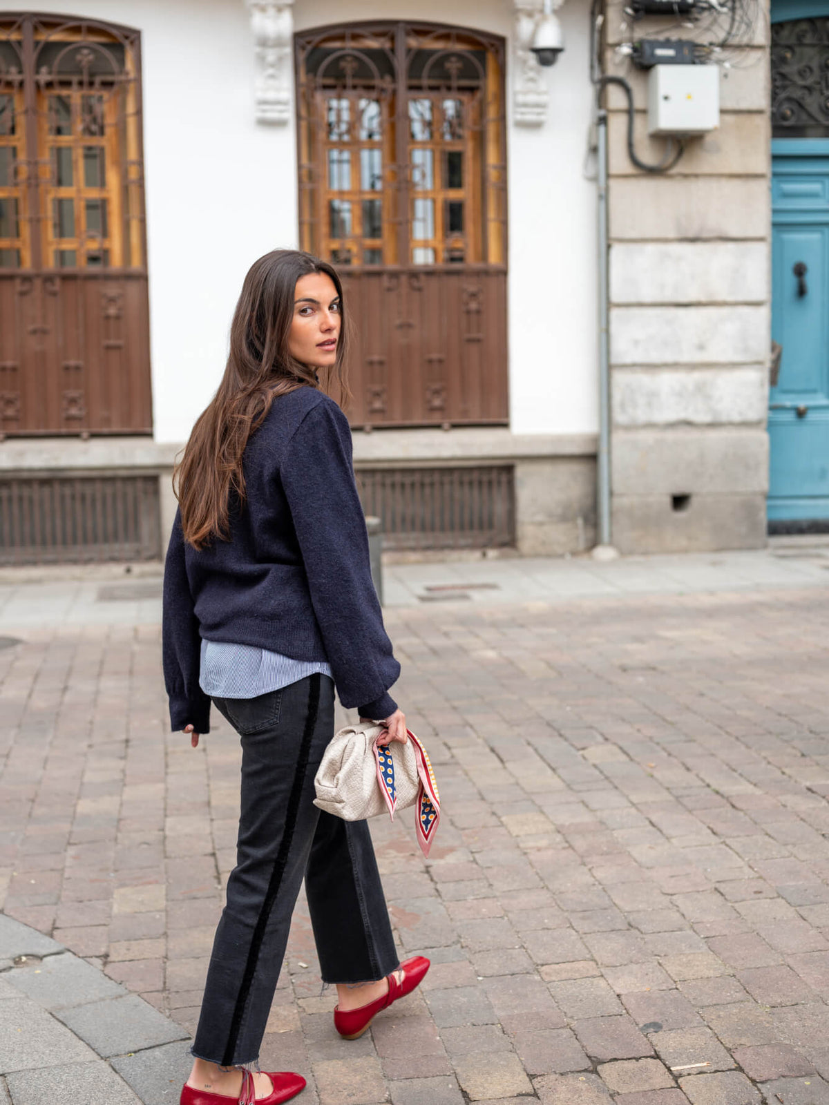 A woman standing on a cobblestone street in front of a building with large windows, wearing a navy blue knit sweater over a grey shirt, black cropped trousers, and red flats, while carrying a small crossbody bag.