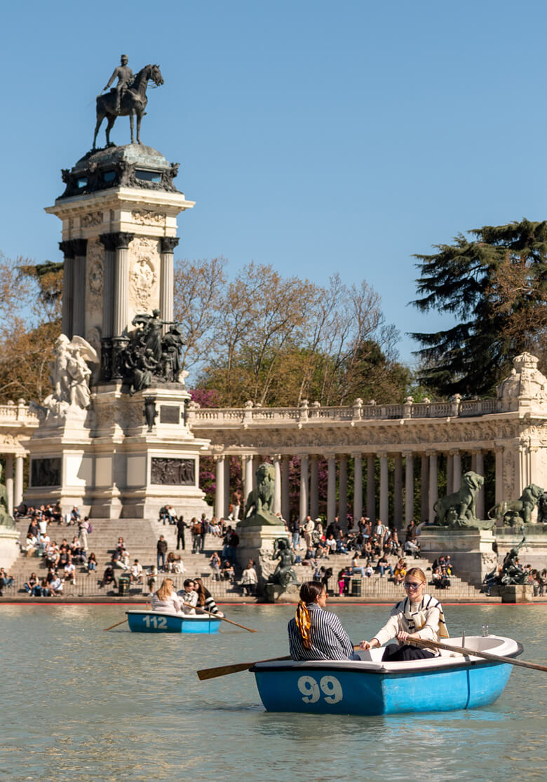 Two people rowing boats on a pond in front of a grand classical stone fountain and monument in Madrid. One boat features the number 112 and the other features the number 99.