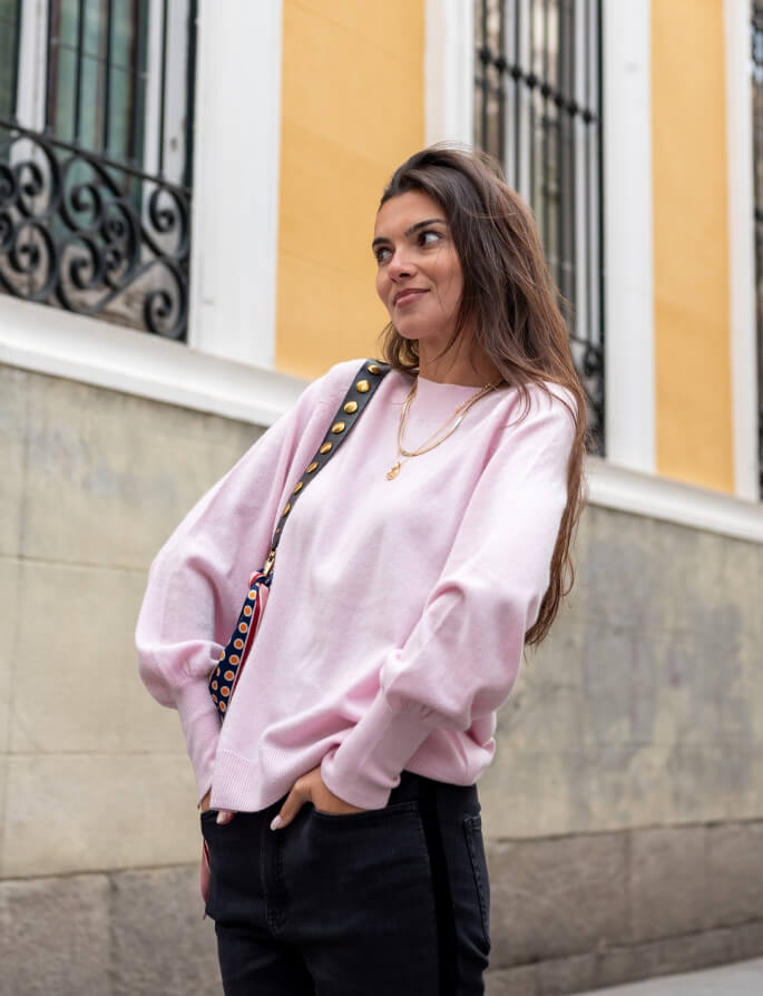 A smiling woman in a soft pink oversized sweater and black trousers standing in front of a yellow building with decorative ironwork in Madrid.
