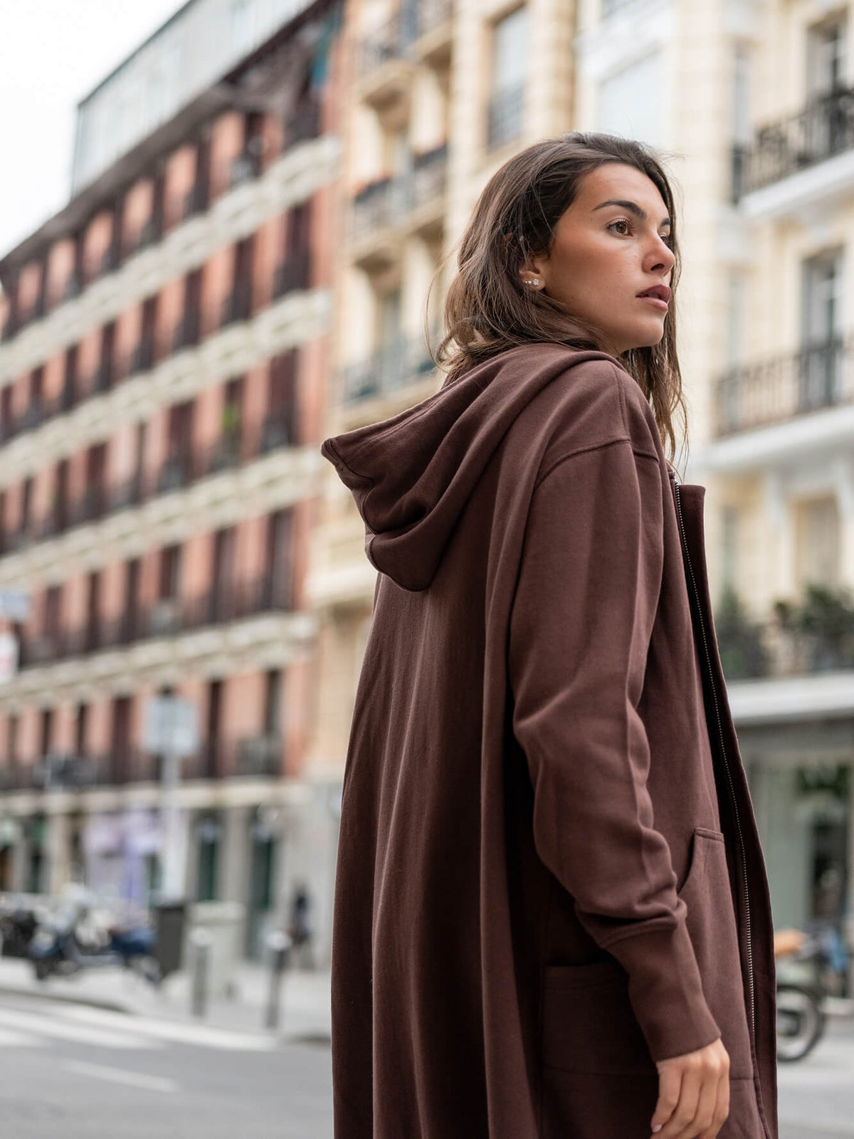 A woman in a cozy, hooded mauve-brown travel-friendly wrap stands on a city street with European architecture in the background.