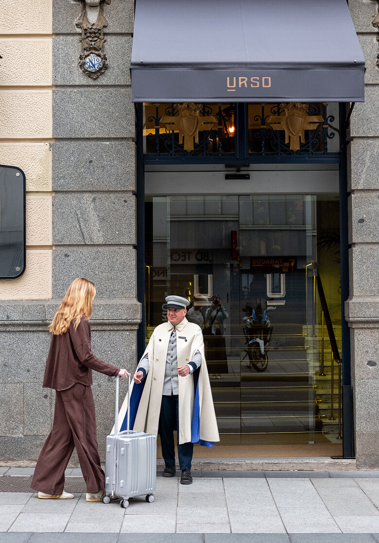 A woman in a brown jumpsuit stands with a suitcase, interacting with a doorman in a white and blue uniform in front of a shop with a gray awning. Text: URSO