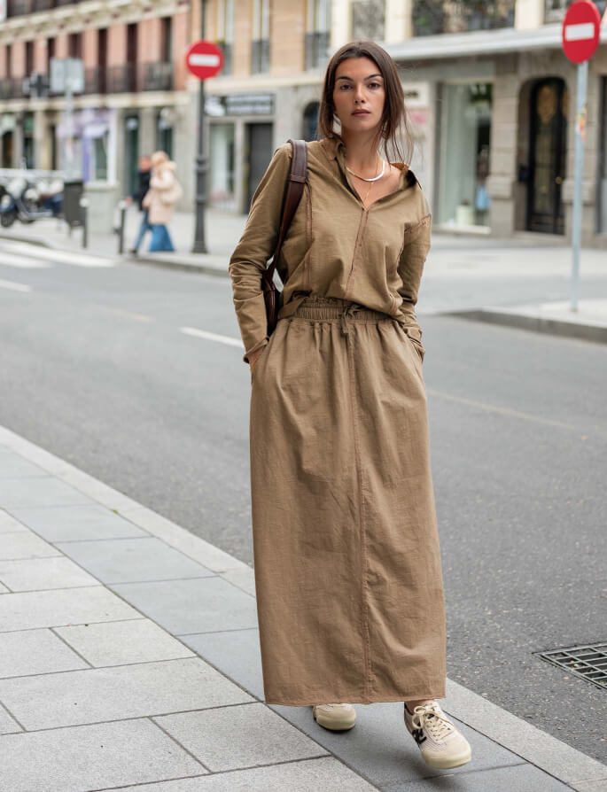 A woman stands on a city sidewalk in front of a building, wearing a long, earthy-toned linen-blend button-down dress and a crossbody bag.
