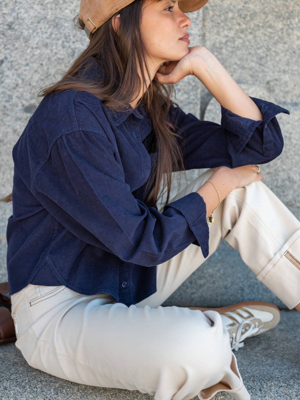 A woman wearing a straw hat and a navy blue button-down shirt sits against a stone wall, wearing cream-colored wide-leg trousers.