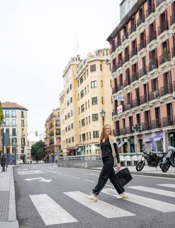 A woman with long blonde hair walking across a city street in Madrid, wearing a black and beige travel-friendly outfit and carrying a large black tote bag, with a classic building and a motorcycle in the background.