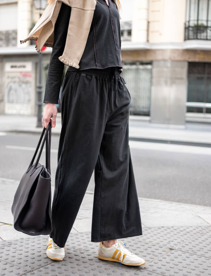 A woman walking down a city street wearing a black knit top, oversized black trousers, and a tan travel wrap, while carrying a large black tote bag.