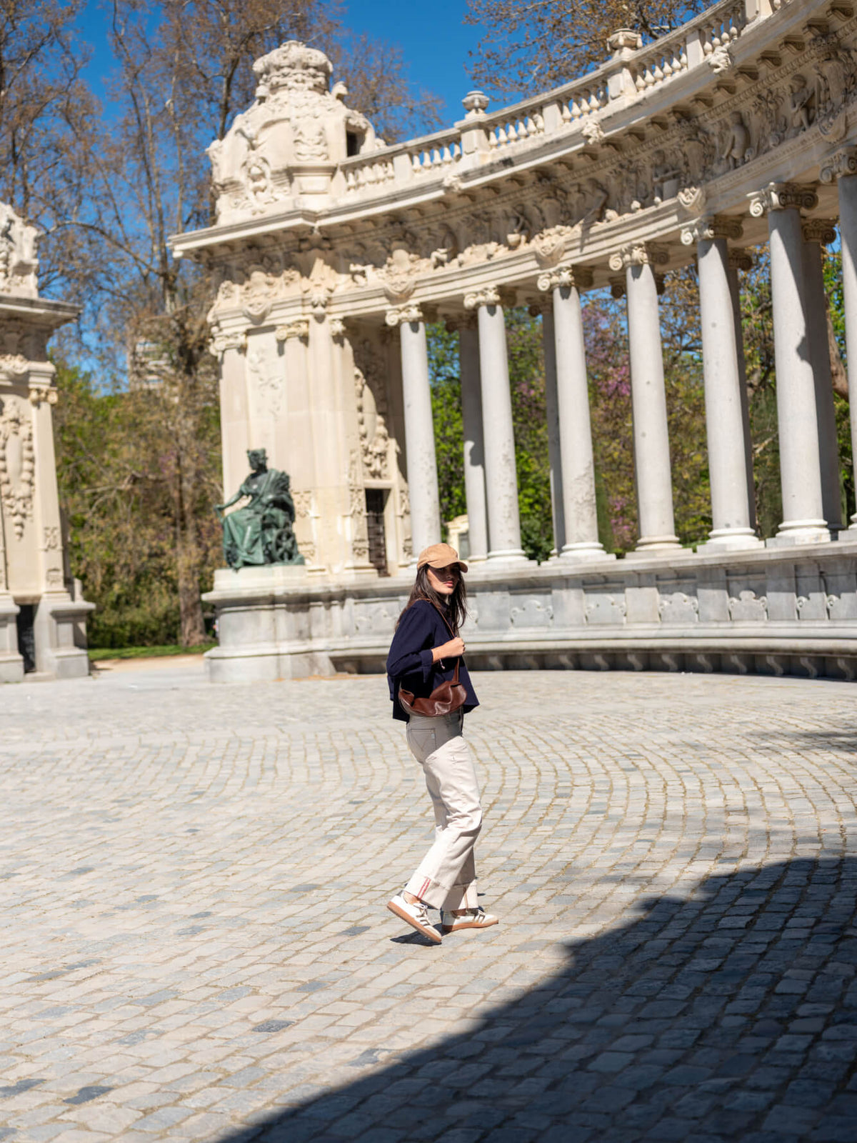 A woman wearing a navy blue top and light-colored trousers stands in a sunlit plaza in front of a large, ornate stone archway and a series of classical columns.