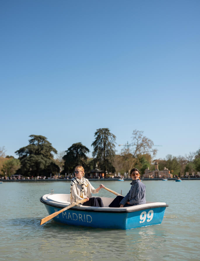 Two women smiling and sitting in a small blue boat on a calm lake with a park and trees in the background. One woman wears a cream knit wrap and the other wears a blue and white striped shirt. The boat has 'MADRID' and '99' printed on the side.