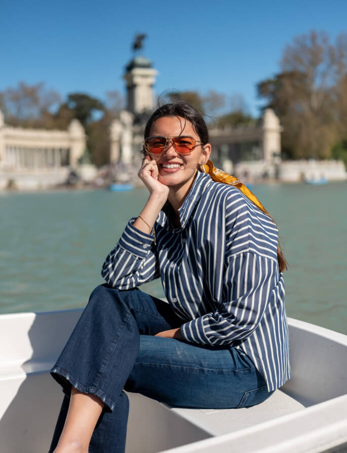 A smiling woman wearing orange sunglasses and a navy and white striped button-down shirt with a yellow silk scarf, sitting on a white boat in a lake with a historic palace and gardens in the background.