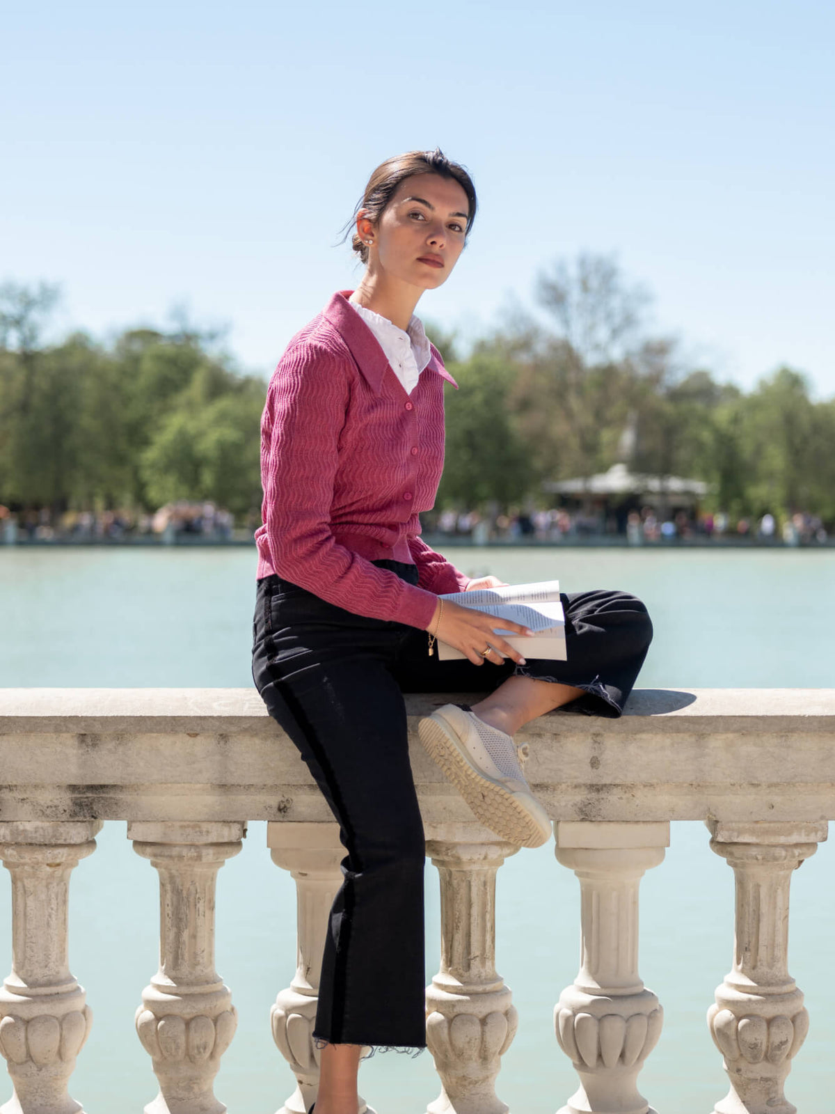 A woman in a pink ribbed knit top and black trousers sits on a stone ledge by a lake, holding a book in a sunny outdoor park setting.