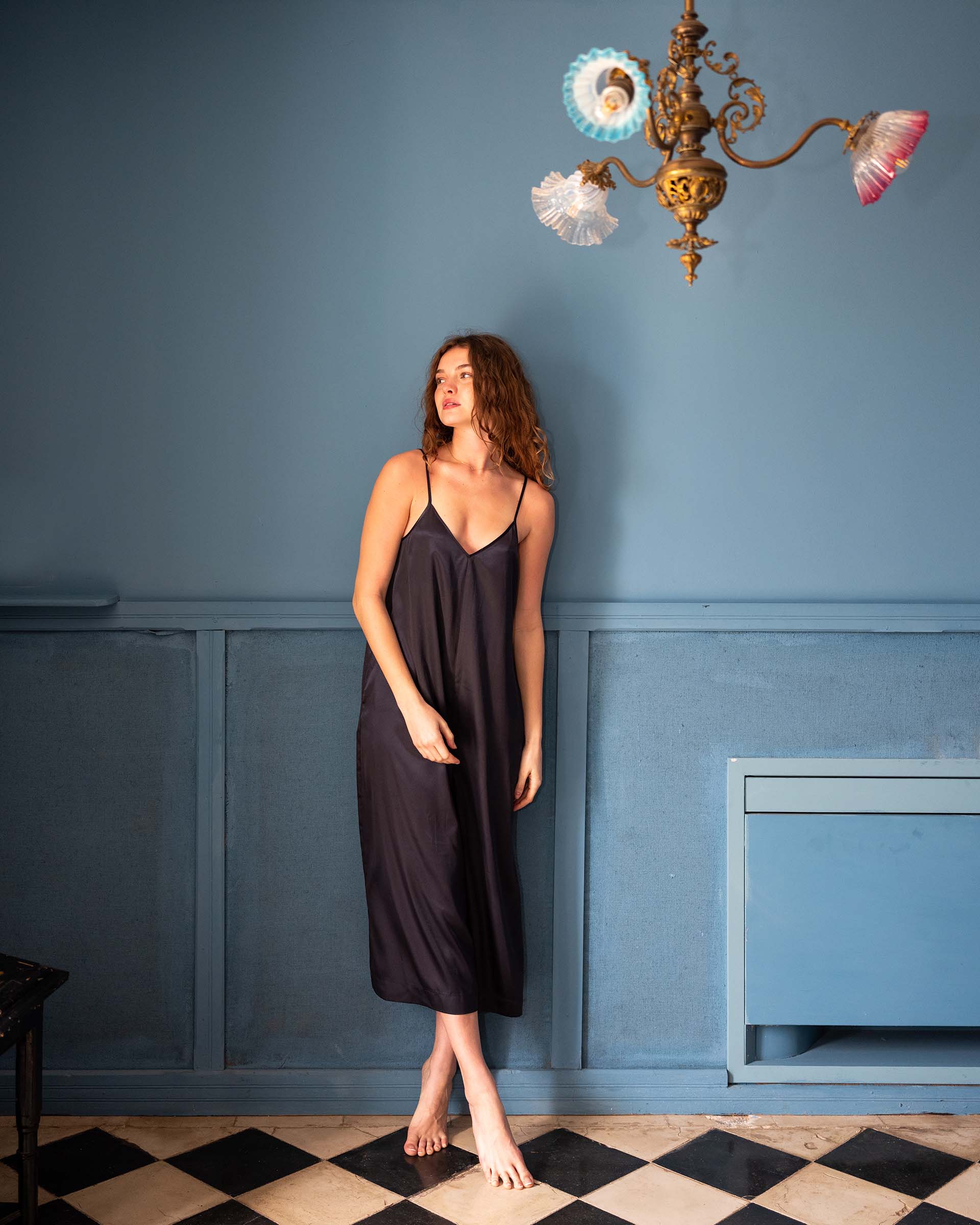 female wearing blue satin jumpsuit standing in front of a wall barefoot