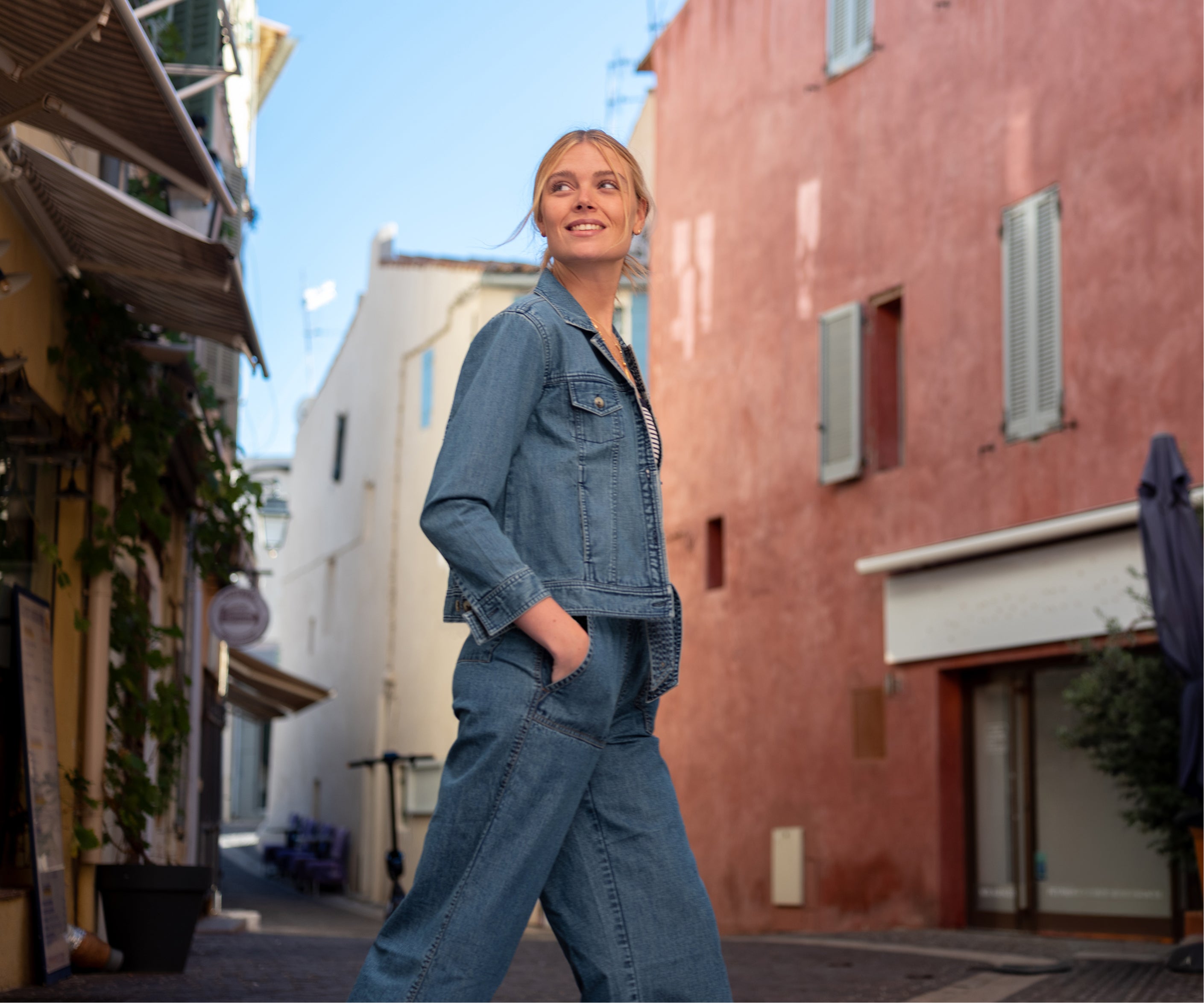 Woman in a denim outfit walking down a sunlit street with buildings on either side.