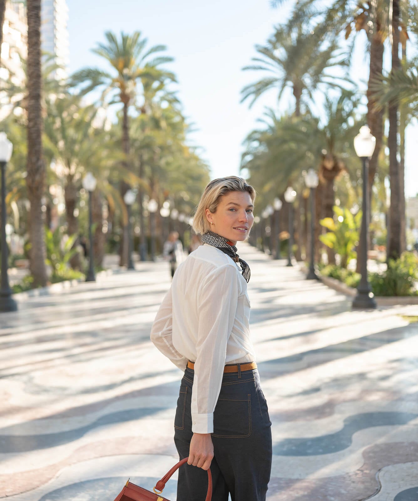 A woman in a white button-down shirt and dark trousers stands on a palm-lined promenade under clear skies.