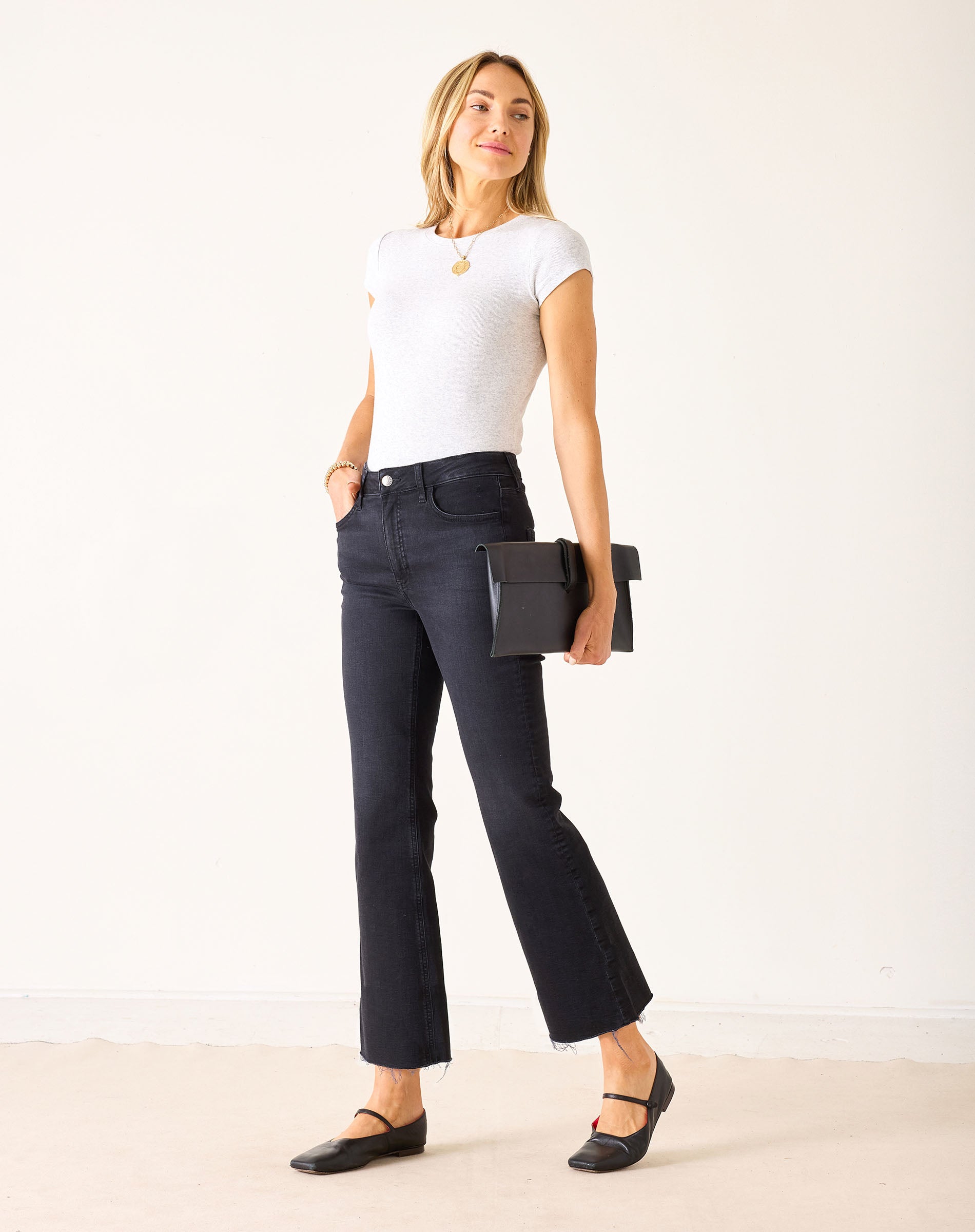 Woman wearing a white t-shirt and black jeans holding a black clutch against a white background