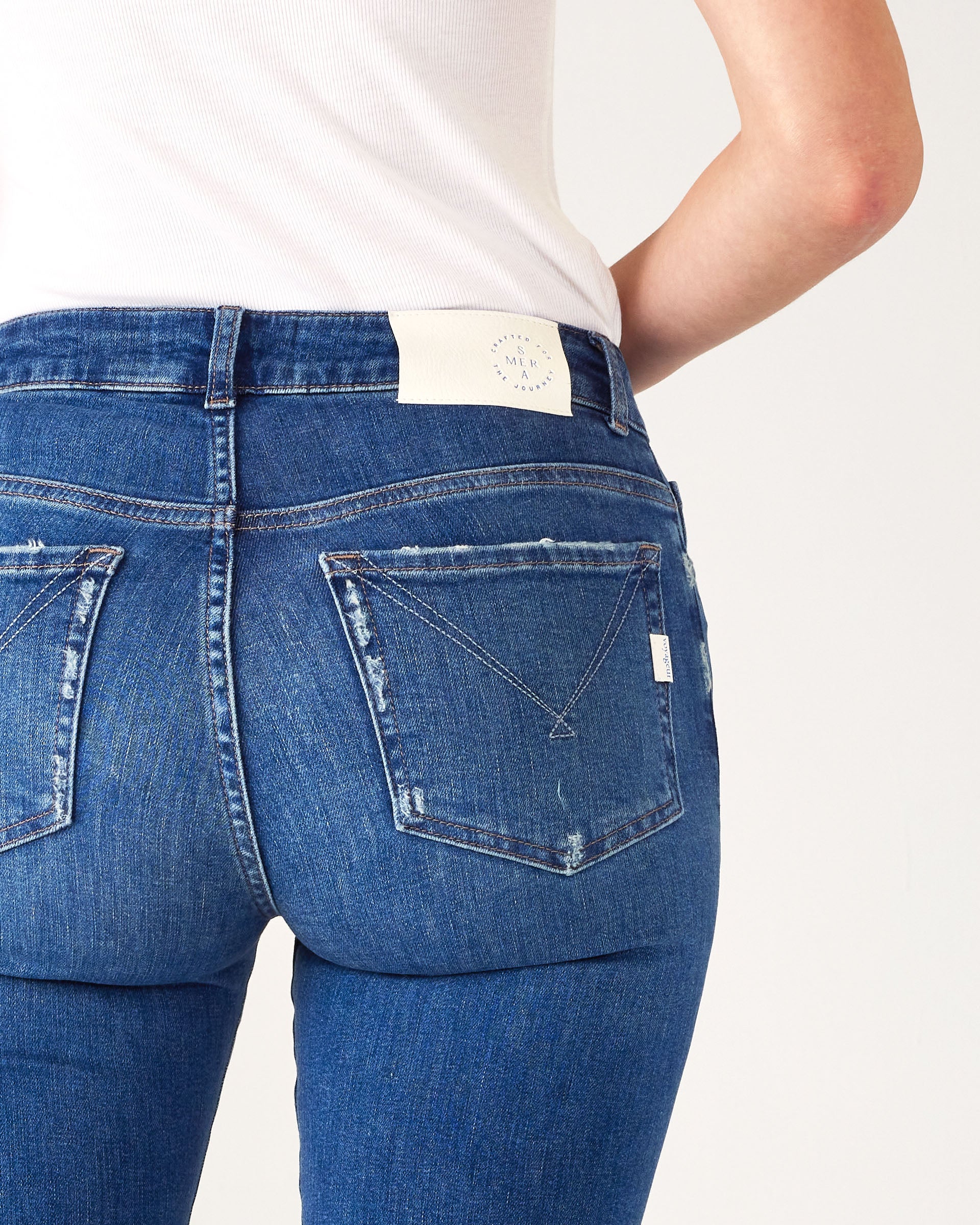 Closeup rear view of Woman wearing Mersea Infinity blue Nomad cropped mini boot-cut jeans standing against white background