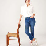 Woman wearing Mersea Infinity blue Nomad cropped mini boot-cut jeans standing with arm on chair against white background