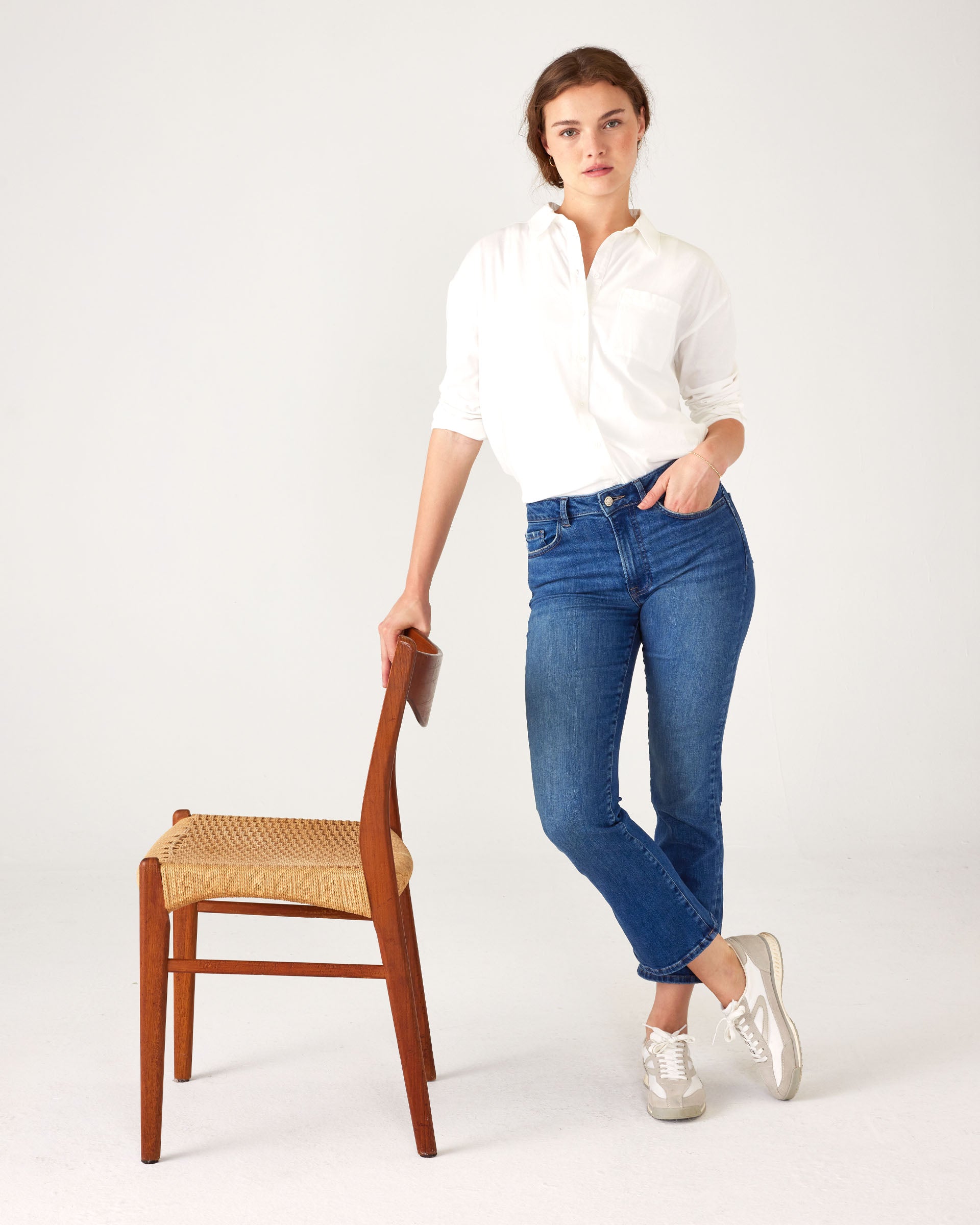Woman wearing Mersea Infinity blue Nomad cropped mini boot-cut jeans standing with arm on chair against white background