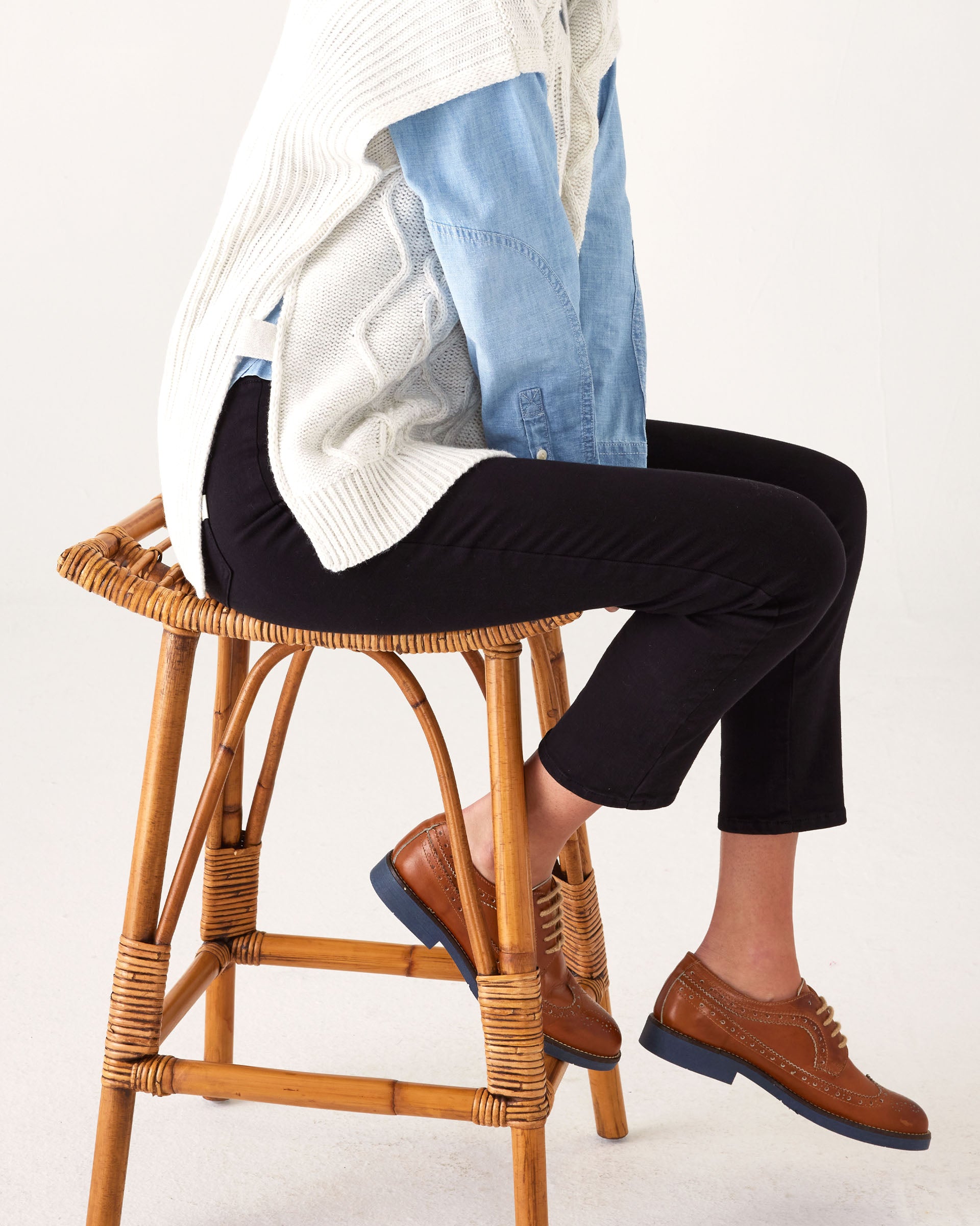 Close-up of lower body: Woman wearing Mersea Nomad black ankle cigarette jeans, sitting on stool against a white background