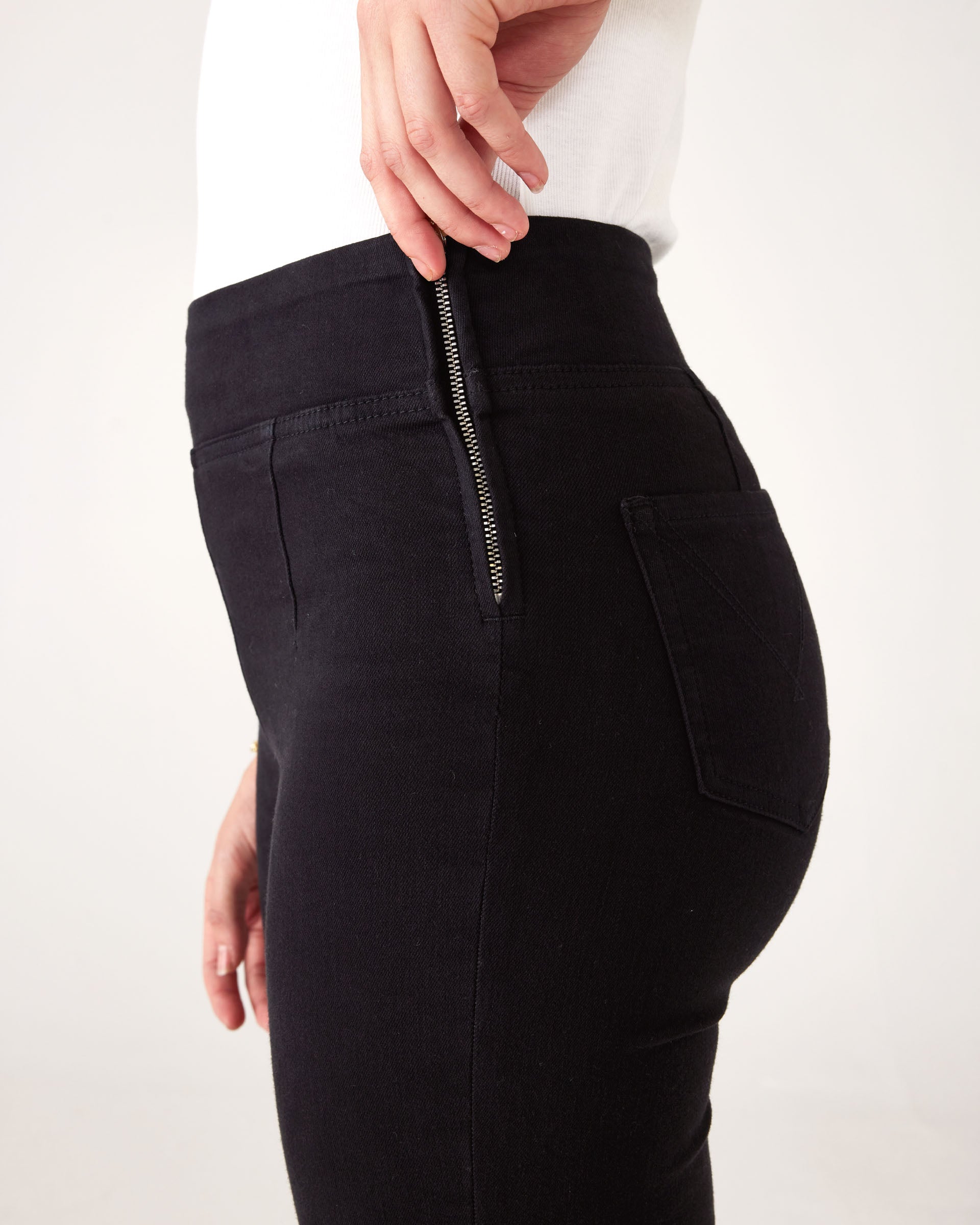 Close-up of side zipper: Woman wearing Mersea Nomad black ankle cigarette jeans, standing against a white background