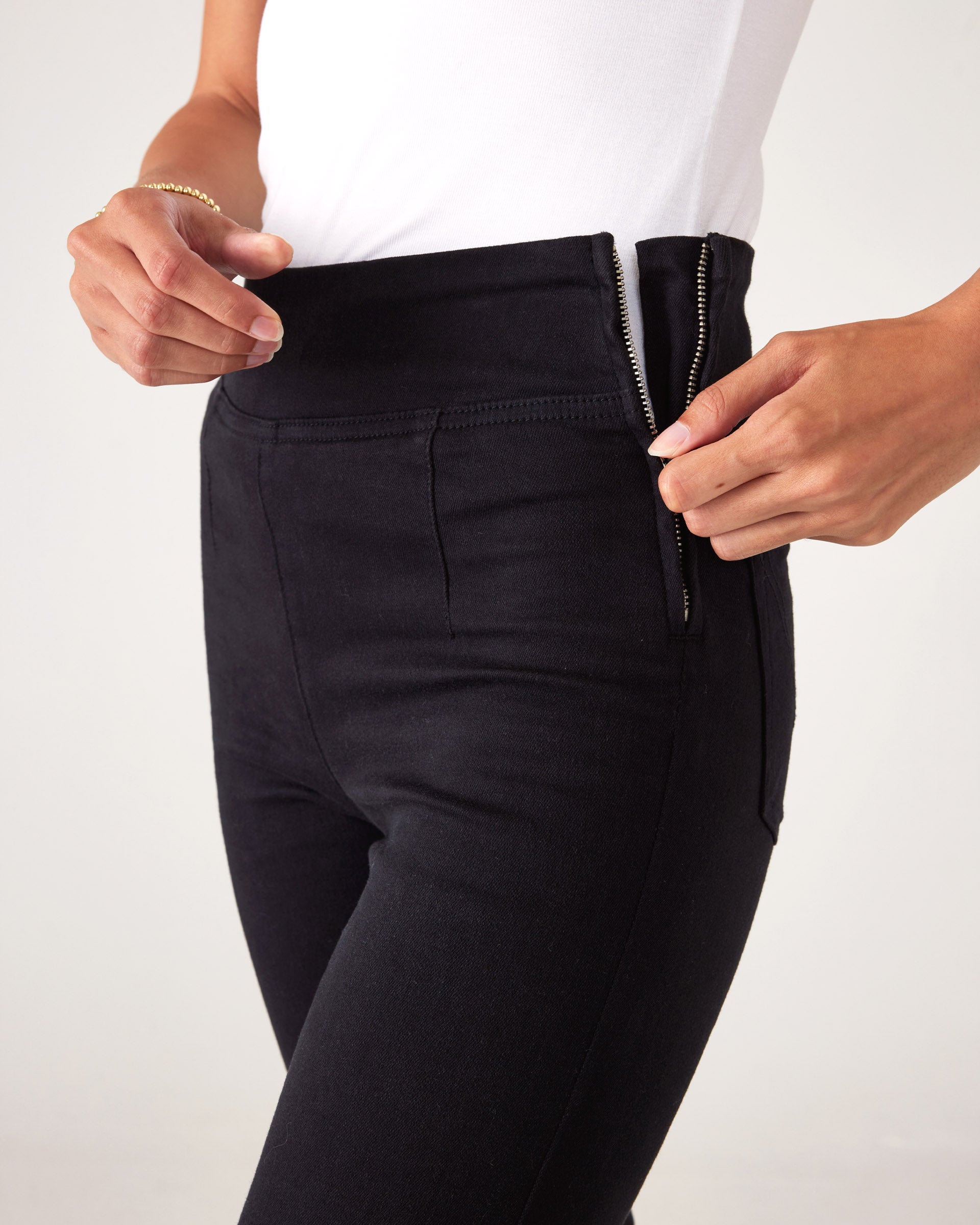closeup view of side zipper on Woman wearing Mersea Nomad black denim full length boot-cut jeans against a white background
