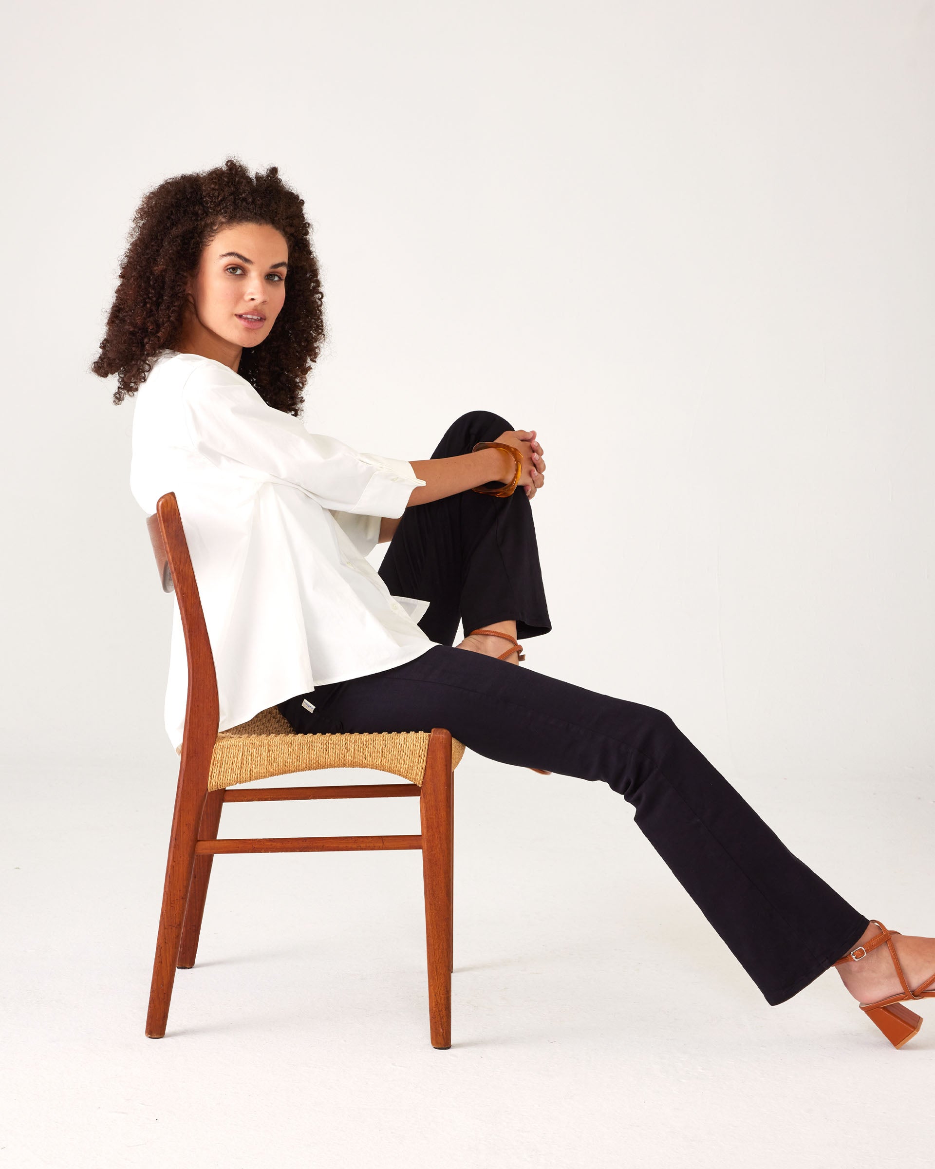 Woman showcasing Mersea Nomad black denim full length boot-cut jeans sitting in a chair against a white background