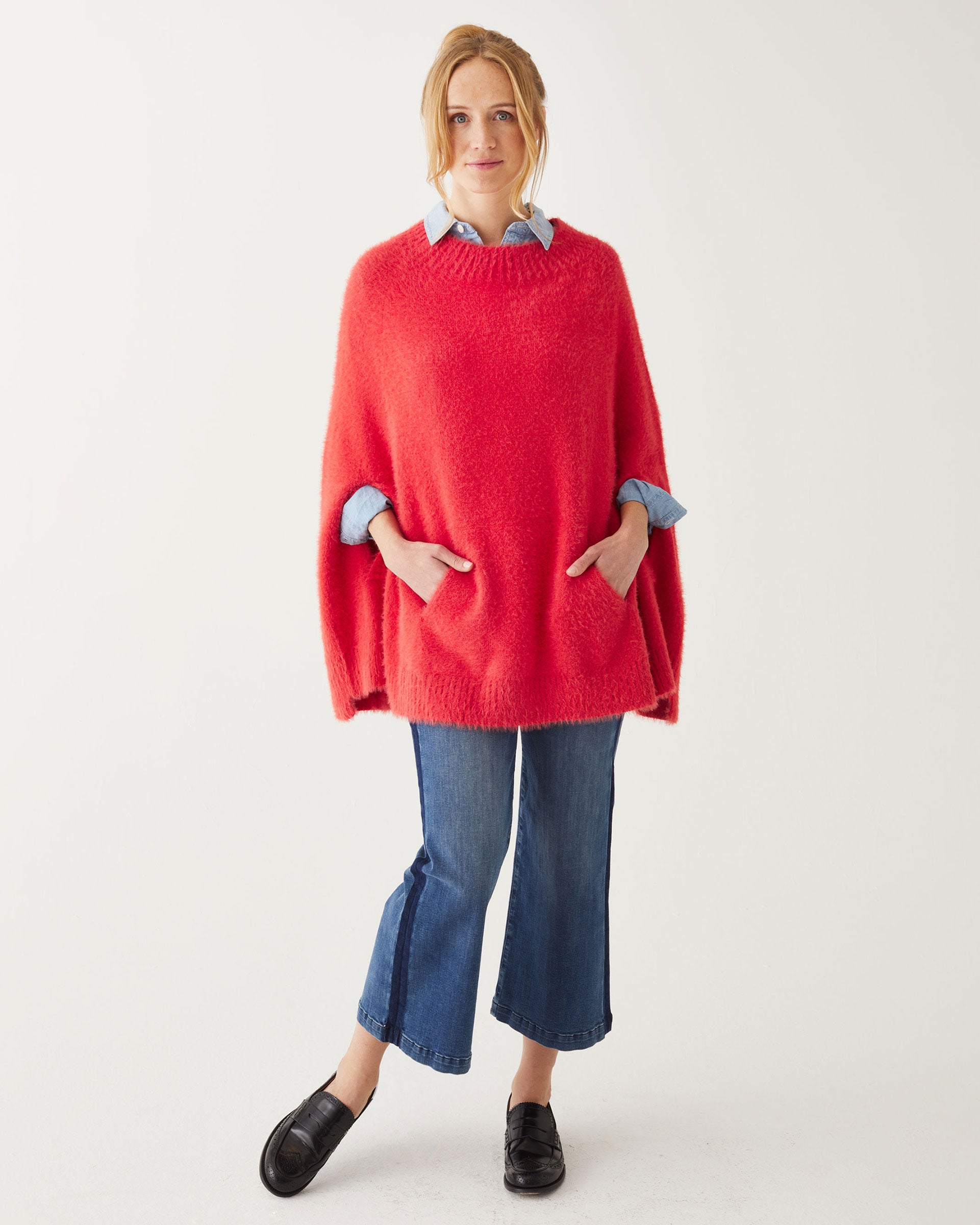 Female wearing a red poncho over blue jeans has her hands in pockets in front of white wall