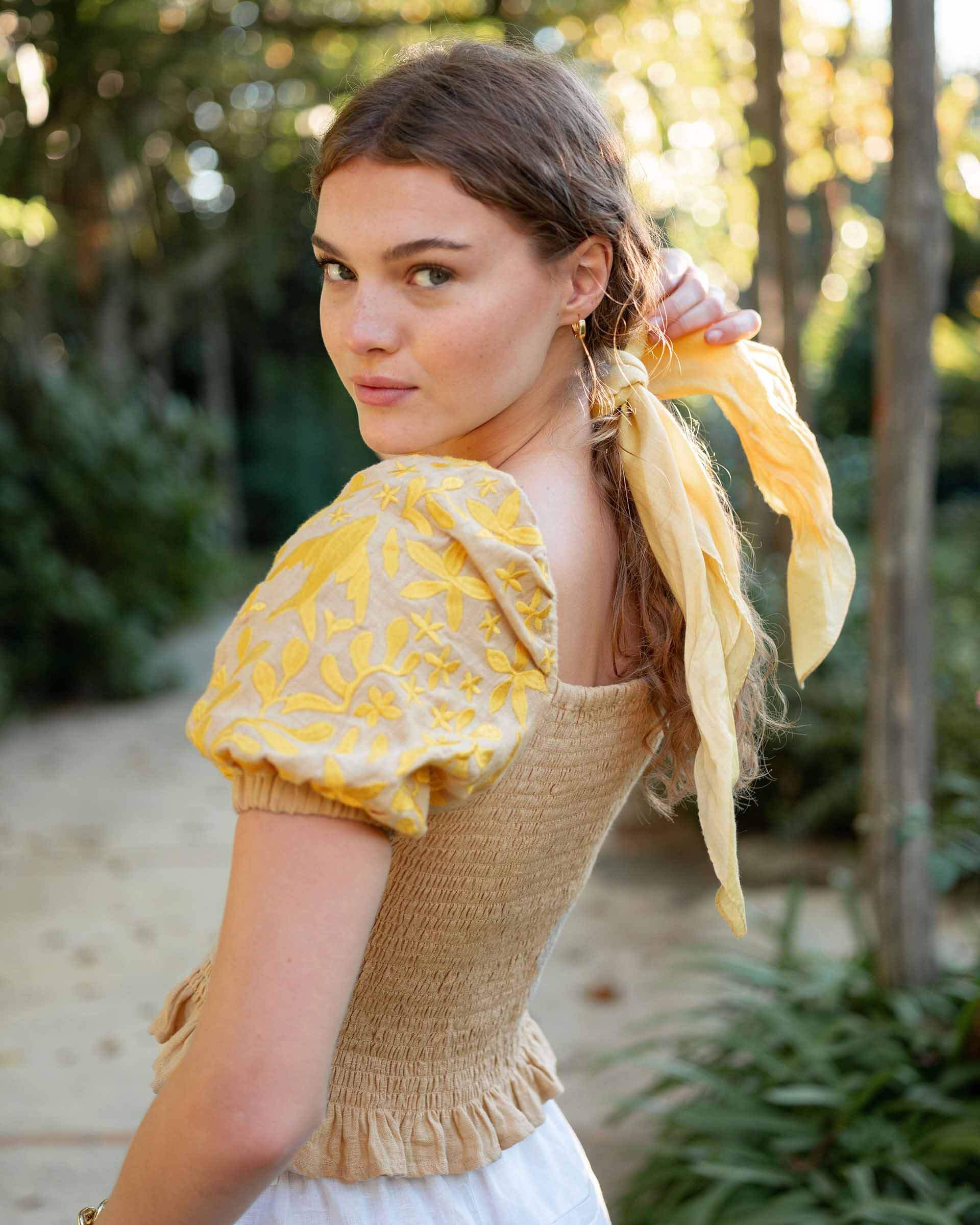 female wearing tan smocked top with yellow embroidery standing outside near trees