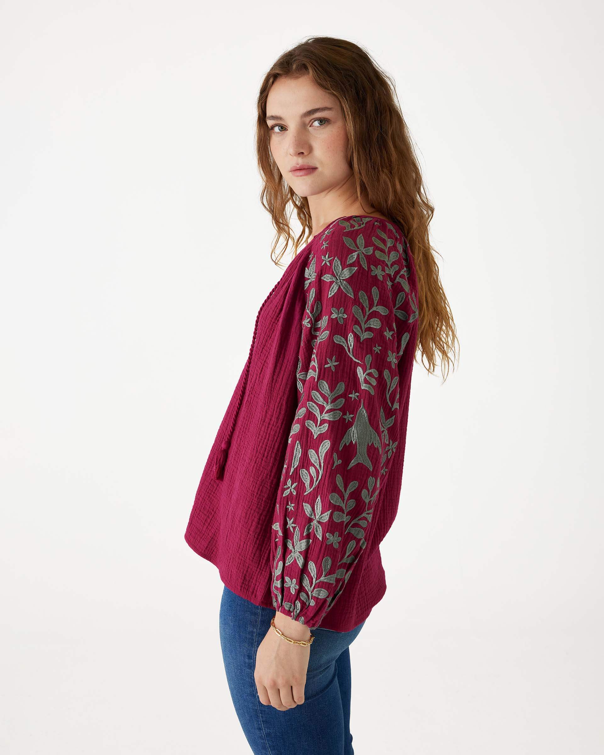 female wearing Mersea berry v-neckline blouse with embroidered sleeves and tassel ties standing sideways on white background