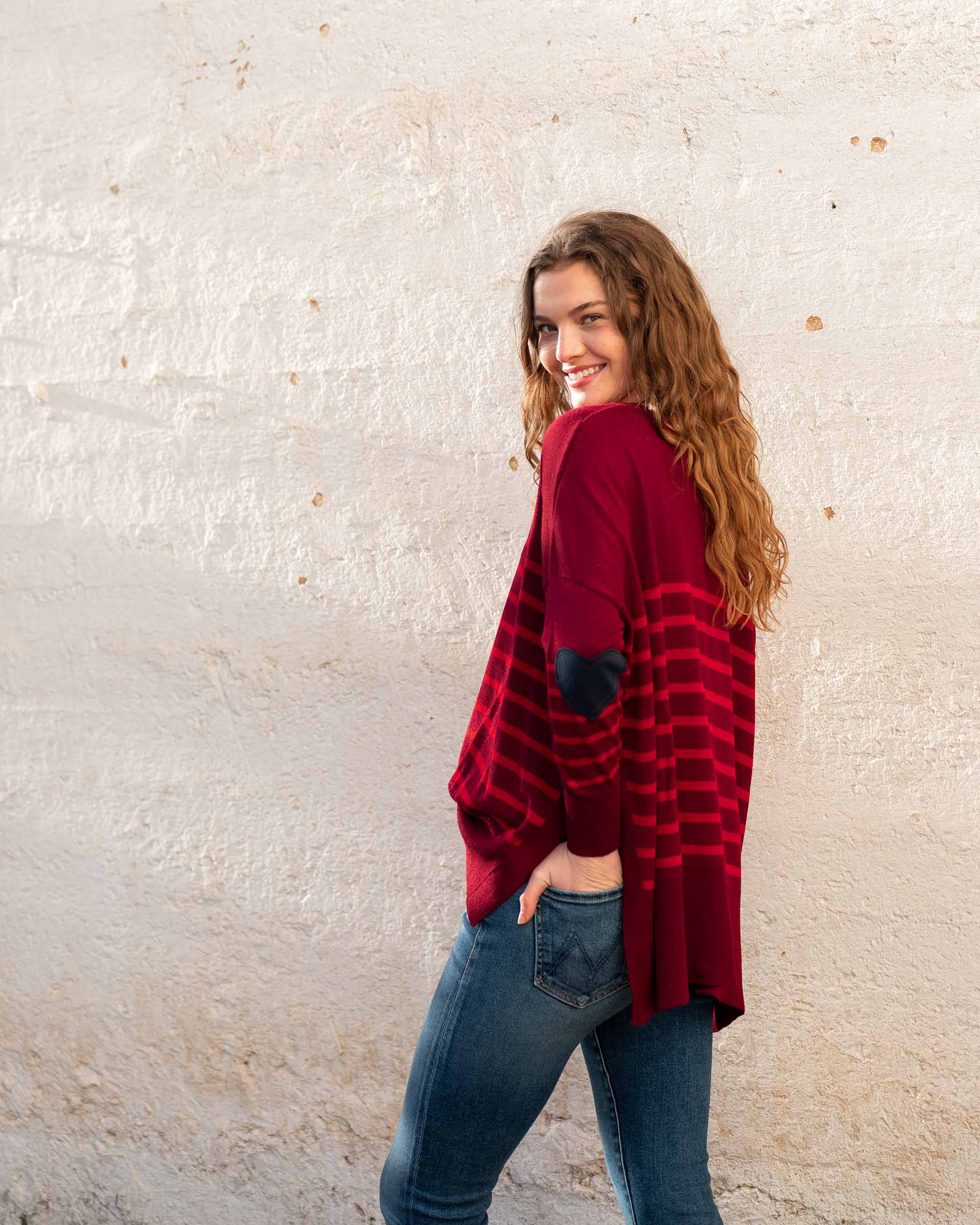 female wearing blue jeans red striped sweater with navy heart elbow patch on a white wall