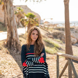 female wearing navy sweater with white stripes and red heart elbow patch standing near a beach