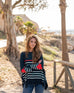 female wearing navy sweater with white stripes and red heart elbow patch standing near a beach
