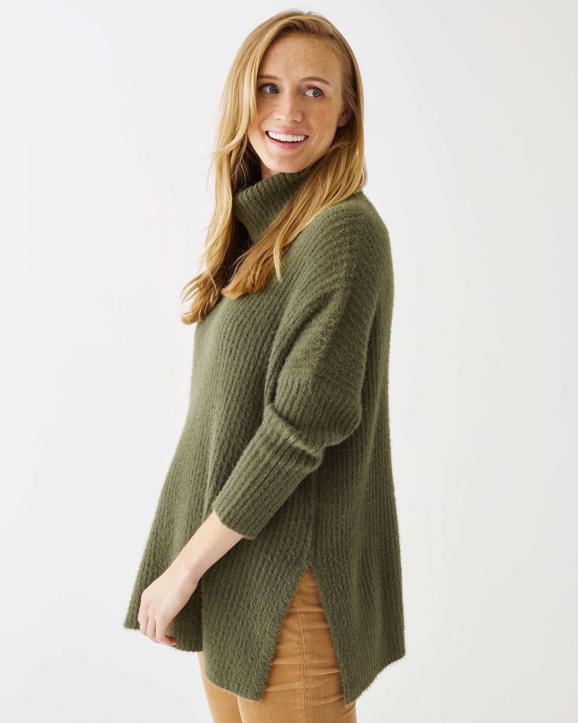 Female wearing a olive green turtleneck sweater with split sides in front of white wall