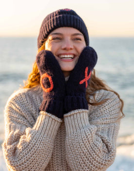 female wearing navy heart beanie with matching XO mittens and neutral sweater on the beach