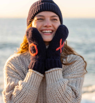 female wearing navy heart beanie with matching XO mittens and neutral sweater on the beach