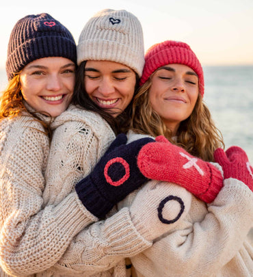 three females wearing matching beanies and mittens hugging on the beach wearing neutral sweaters