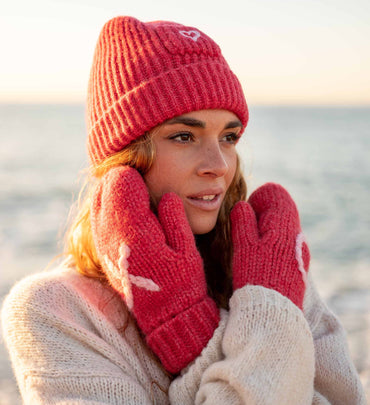 female wearing red heart beanie with matching XO mittens and white sweater on the beach