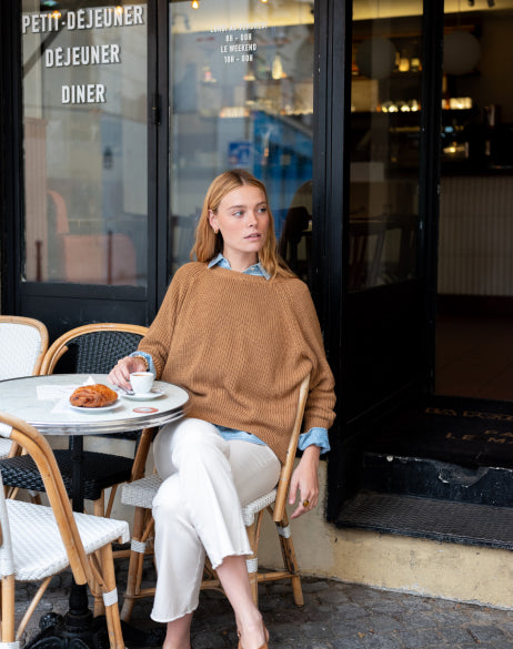 Image of a woman wearing a tobacco knit crewneck sweater sitting at a cafe table.