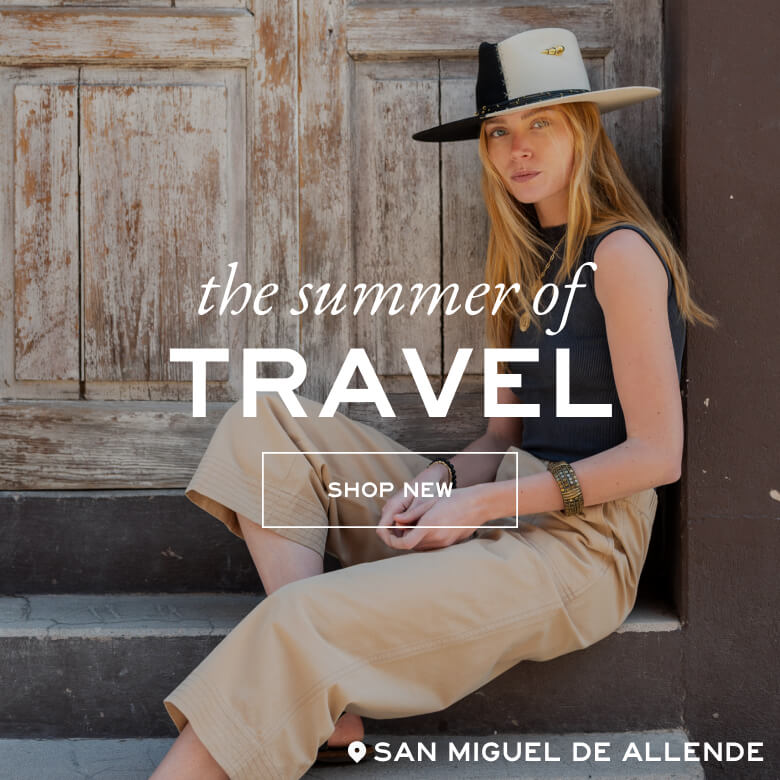 Woman sitting on steps with text overlay 'the summer of travel' and 'Shop New' in front of a rustic wooden door.