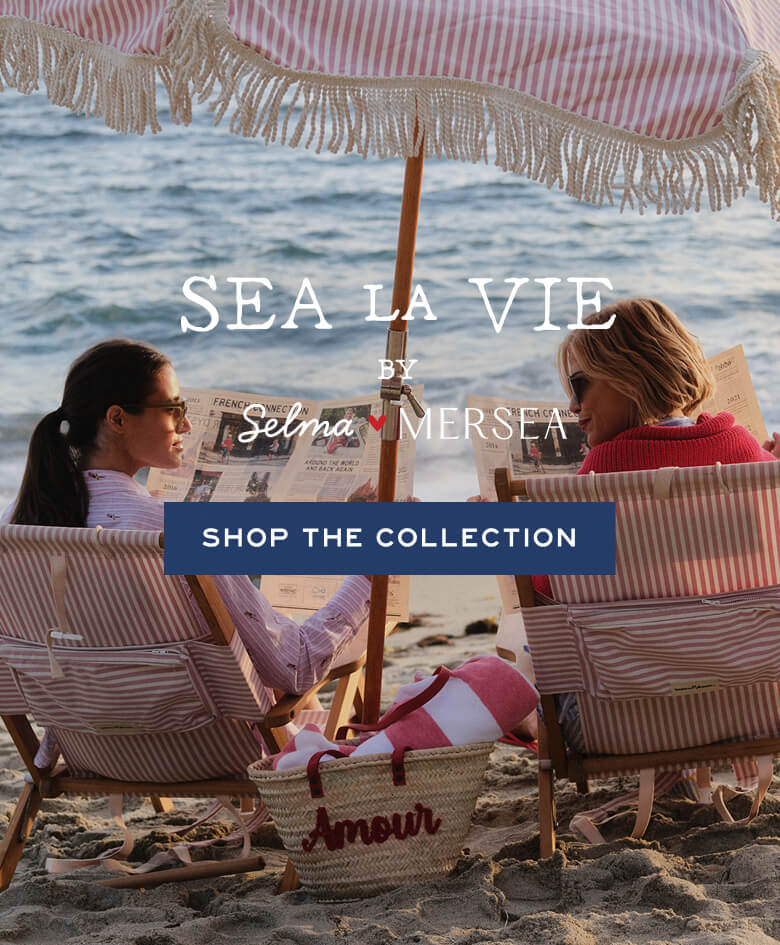 Two women sitting under a striped beach umbrella on the sand with 'Sea La Vie' branding.