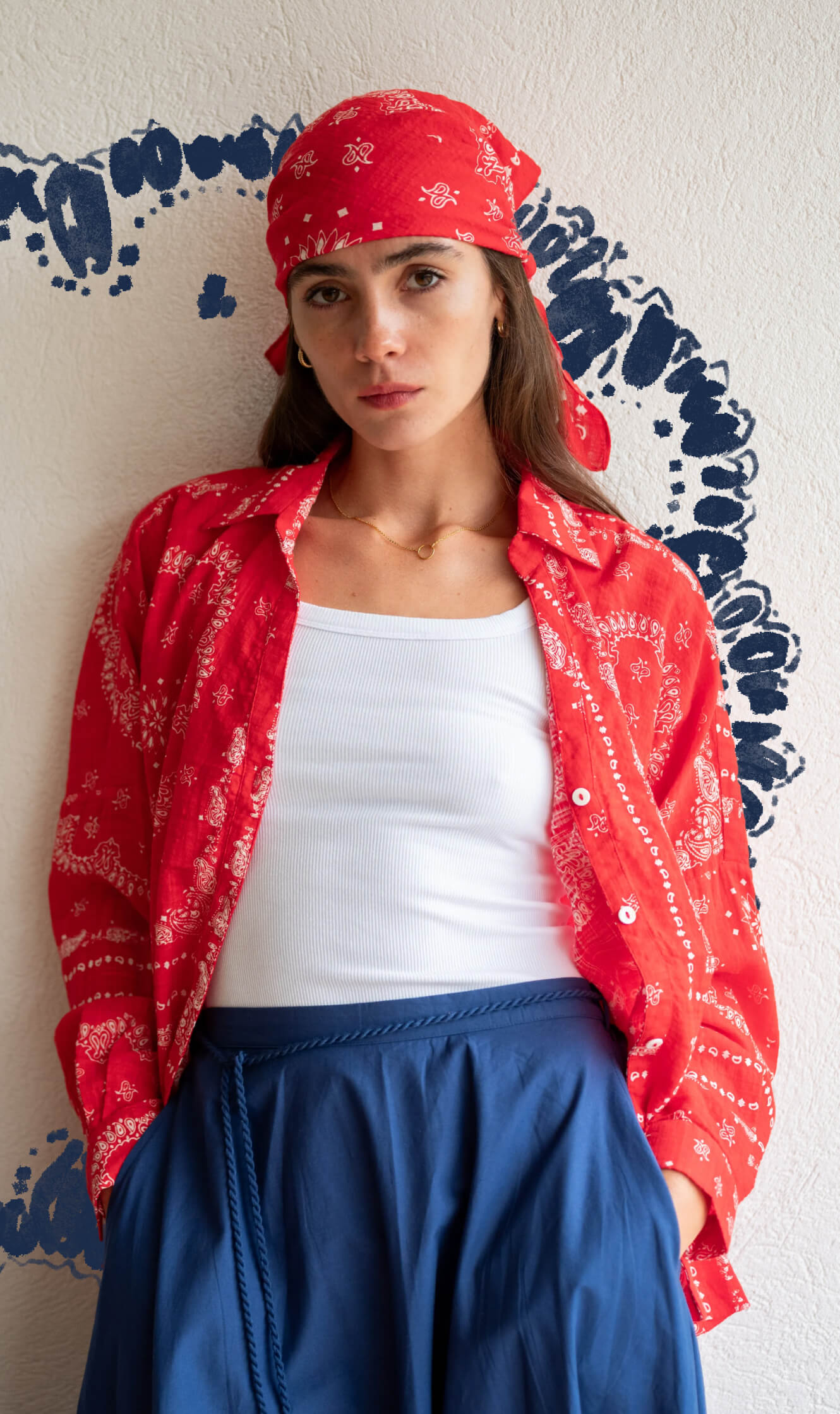 Woman wearing a red bandana, red jacket, white top, and blue pants against a white wall with blue patterns.