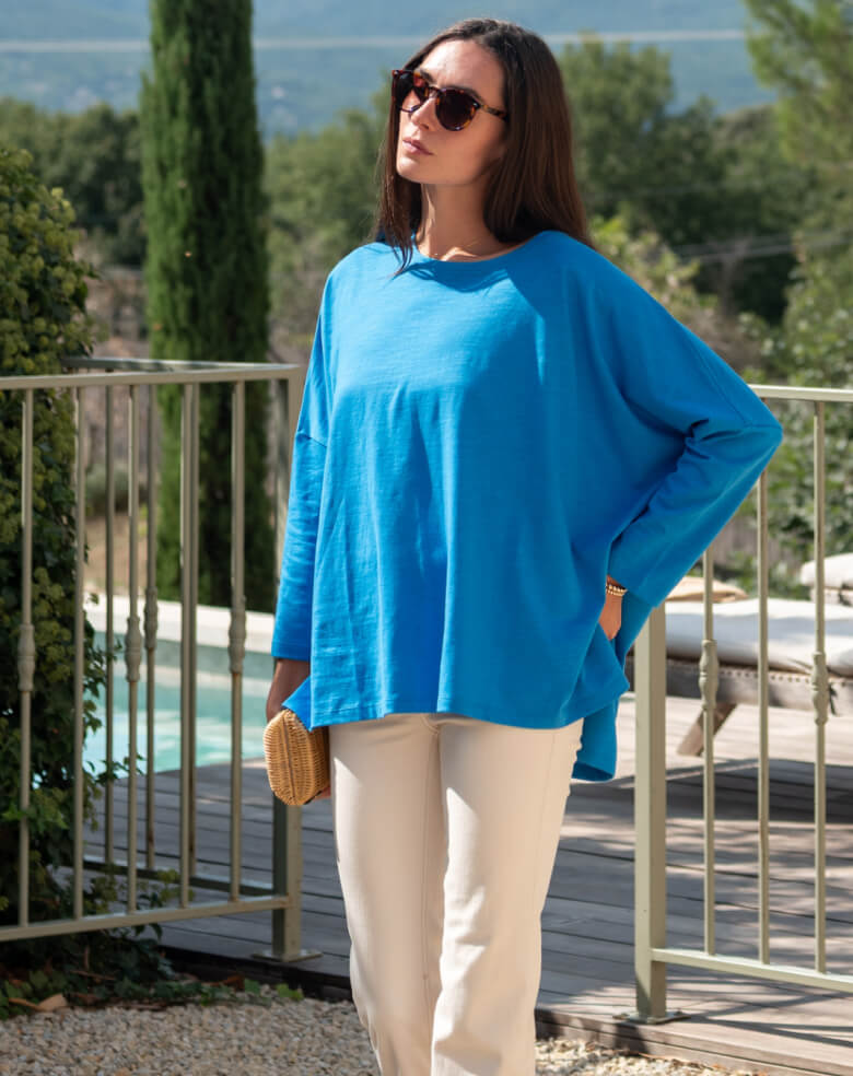 Woman wearing a blue top and white pants standing outdoors with a pool and trees in the background.