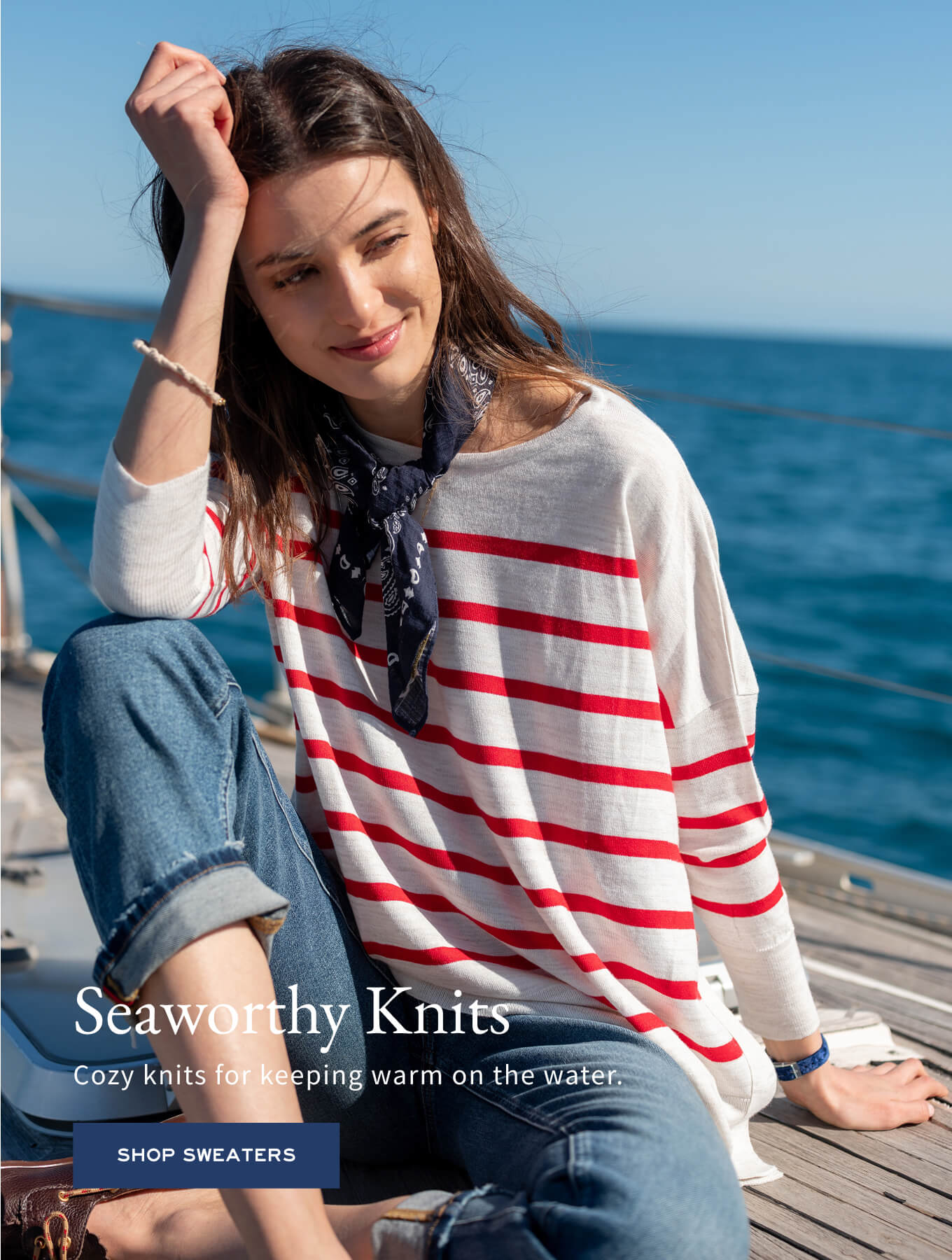 Cream knit sweater with red stripes and a scarf, worn by a model on a boat. Text: Seaworthy Knits Cozy knits for keeping warm on the water. SHOP SWEATERS