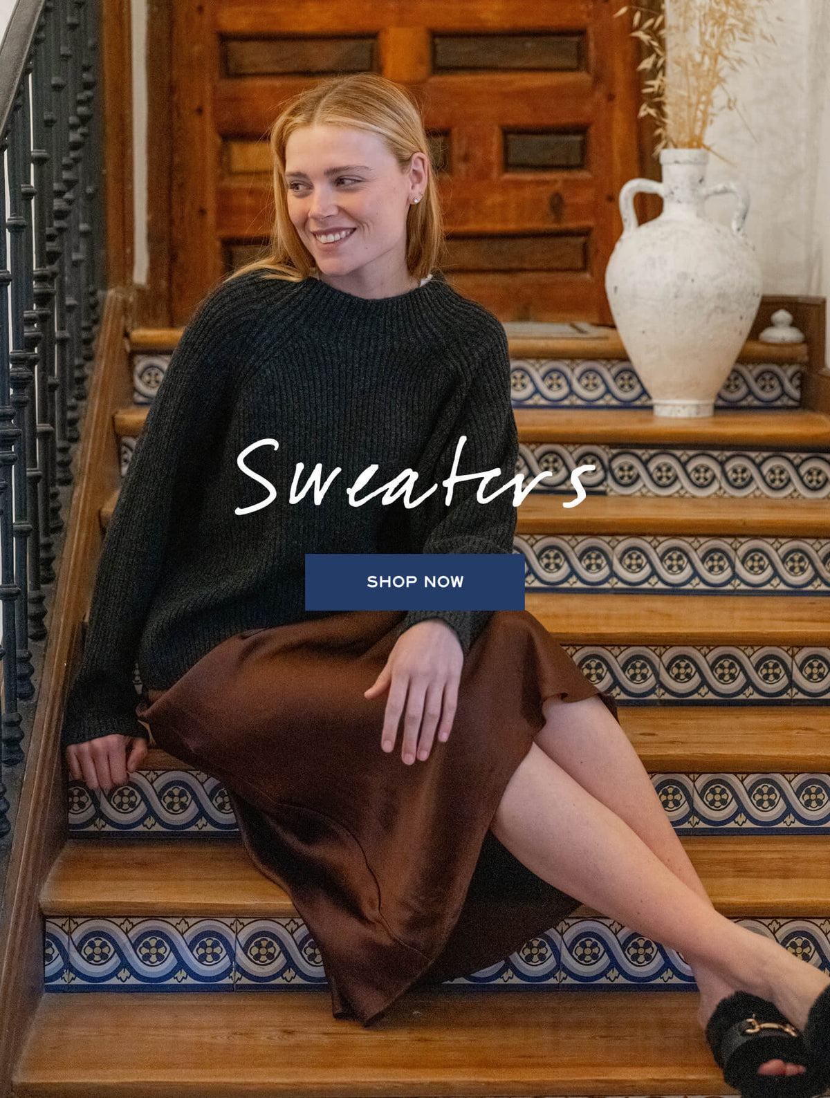 Woman in a knit sweater and satin skirt sitting on a patterned tiled staircase. Text: "Sweaters. SHOP NOW"