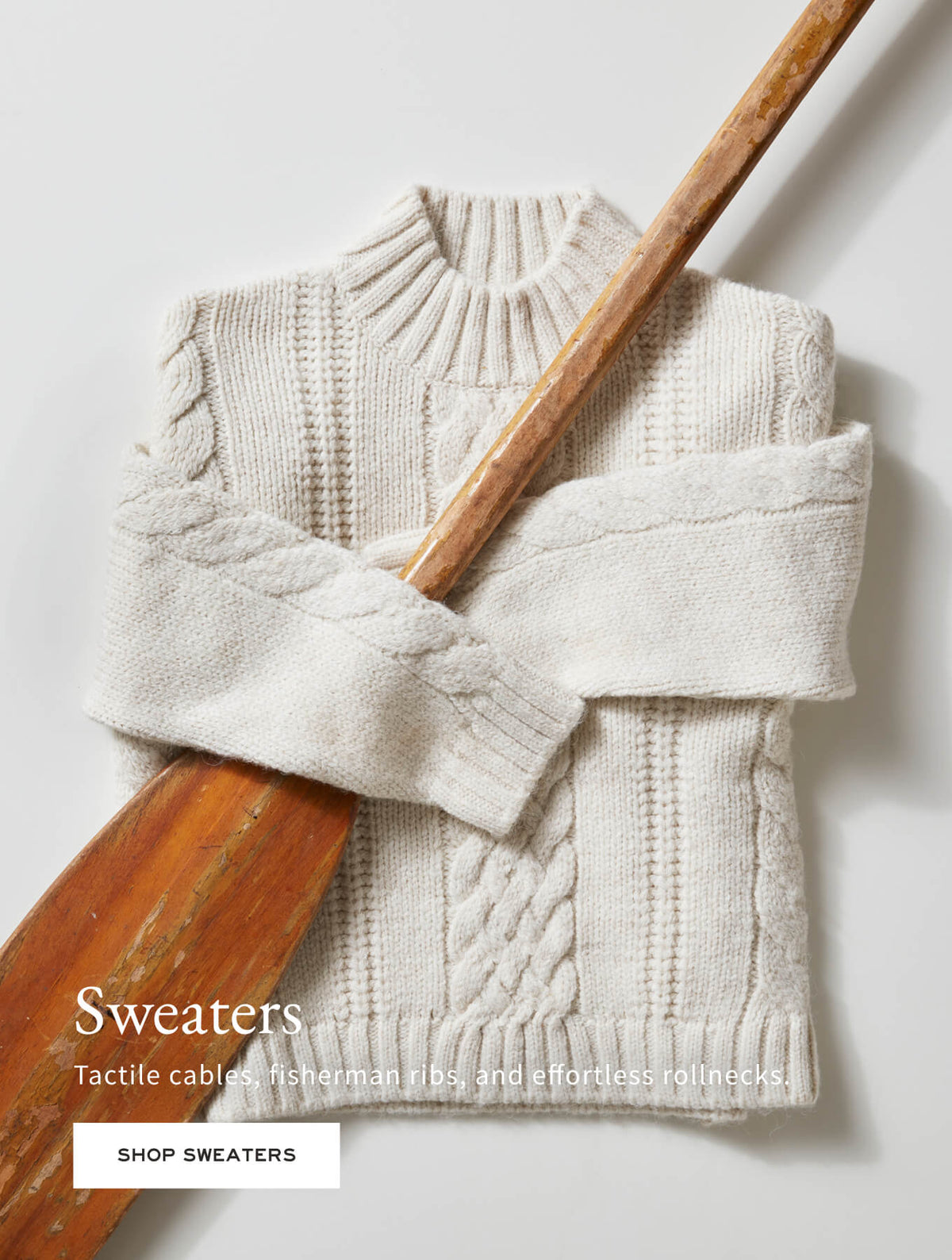 Promotional banner for knitwear. Features a cream-colored cable knit sweater with a roll neck draped over a wooden paddle. Text: 'Sweaters. Tactile cables, fisherman ribs, and effortless rollnecks. SHOP SWEATERS'
