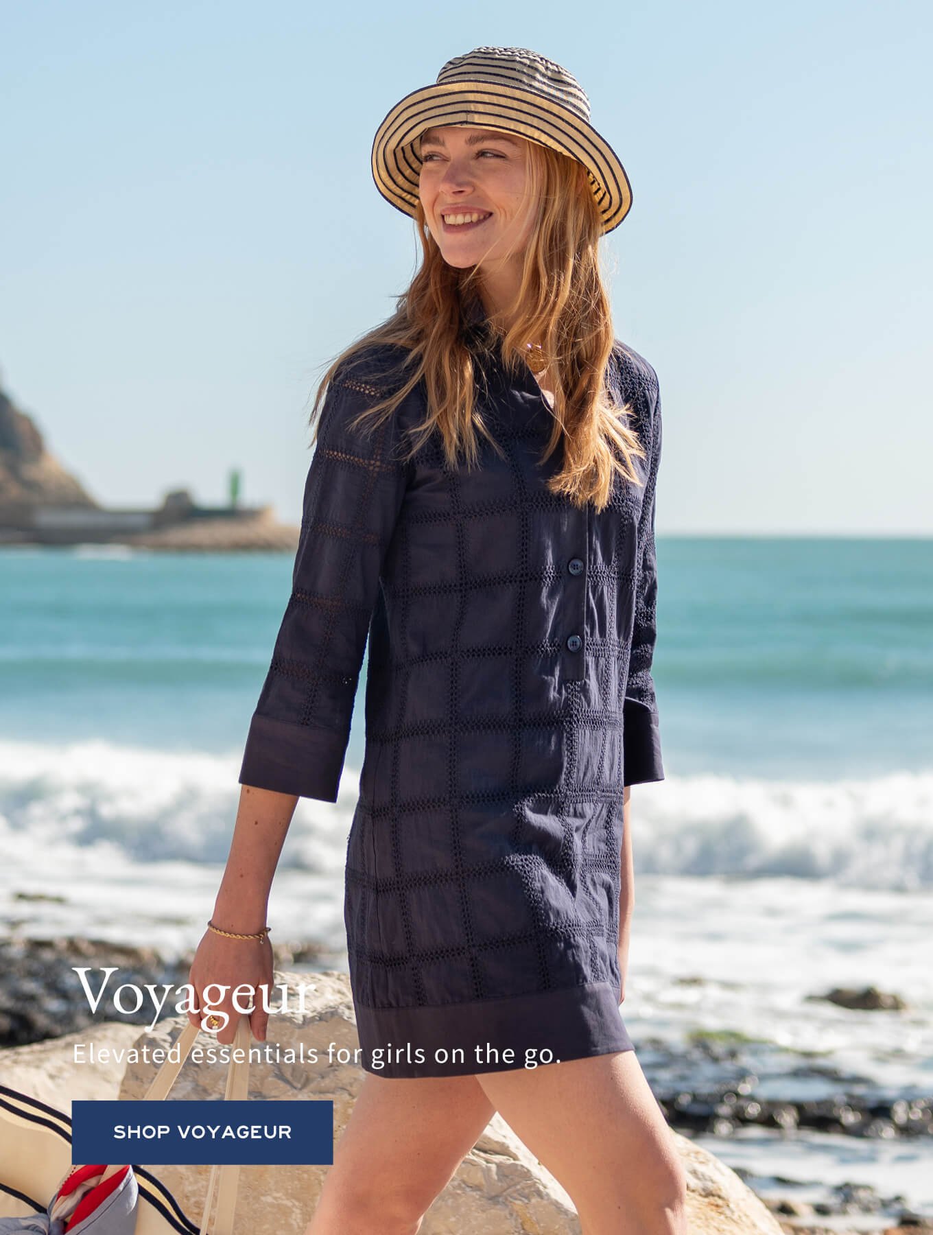Woman in a navy blue textured tunic-style top and straw hat standing on a beach with the text 'Voyageur: Elevated essentials for girls on the go. SHOP VOYAGEUR' superimposed over the image.