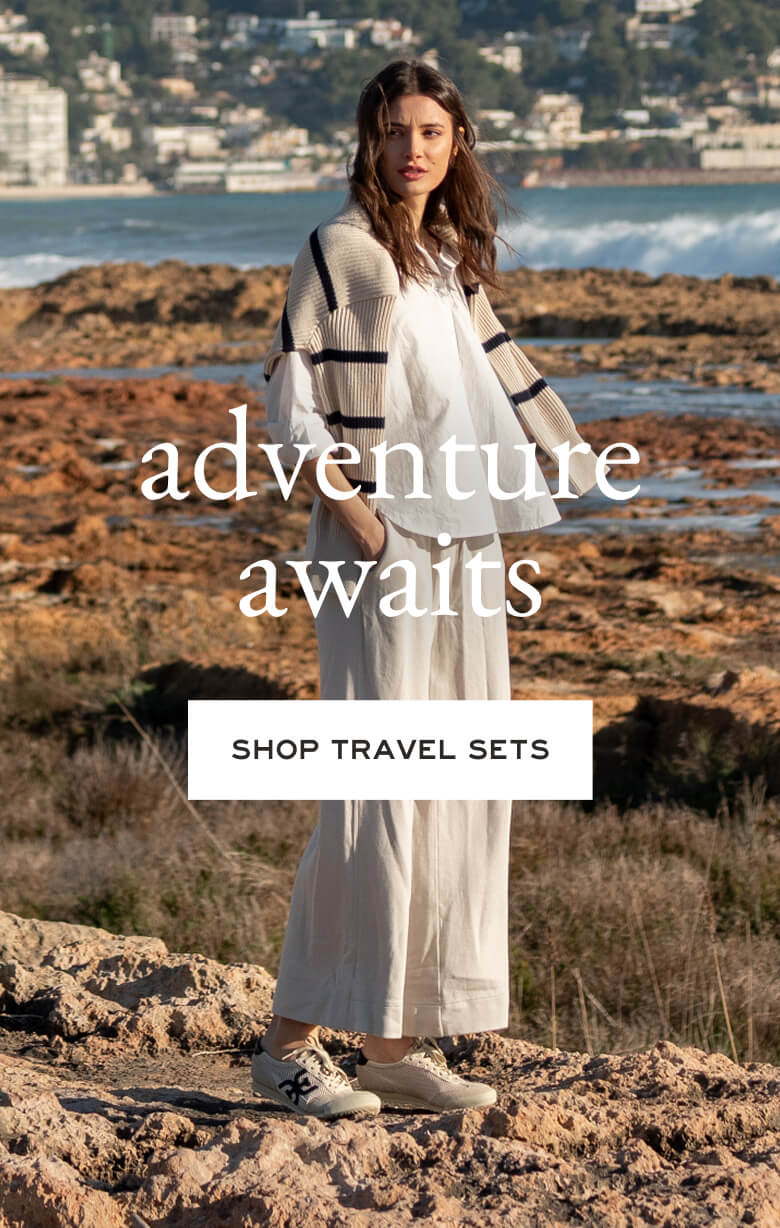 A woman in a travel set standing on a rocky shore near the water. Text: 'adventure awaits', 'SHOP TRAVEL SETS'